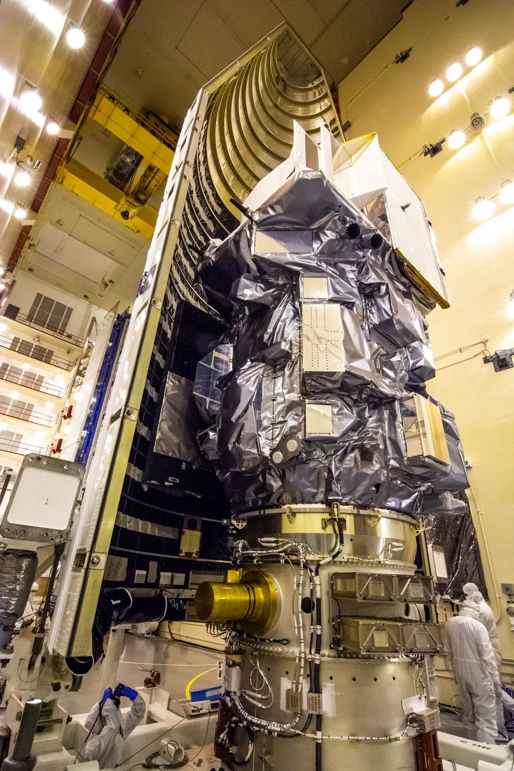 The Landsat 9 spacecraft standing inside a cleanroom, wrapped in silver and gold thermal blanketing, with one half of its launch fairing open beside it and two technicians in white coveralls at its base.